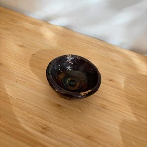 Könnte beinhalten: Eine kleine, dunkelbraune Keramikschale mit glänzendem Innenraum. Die Schale hat eine ovale Form und steht auf einer Holzoberfläche. Der Innenraum hat eine dunkelgrüne und goldene Farbe.