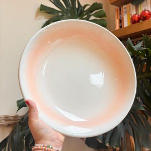 Könnte beinhalten: Eine große, runde weiße Keramikschüssel mit einem sanften, pfirsichfarbenen Farbverlauf am Rand. Die Schüssel wird hochgehalten, wodurch ihr klares Design und ihre glatte Oberfläche zur Geltung kommen. Die Größe der Schüssel deutet darauf hin, dass sie sich zum Servieren von Salaten oder anderen Gerichten eignet.