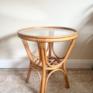 Könnte beinhalten: Ein runder, hellbrauner Beistelltisch aus Rattan mit einer transparenten Glasplatte. Der Tisch hat gebogene Beine und ein dekoratives Webmuster in der Nähe des Sockels. Der Tisch hat einen Durchmesser von ca. 61 cm und ist 51 cm hoch.