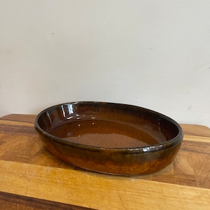 Può includere: Una pirofila ovale in ceramica marrone con finitura lucida. Il piatto ha un bordo leggermente rialzato ed è appoggiato su un tagliere di legno. L'interno del piatto riflette la luce.