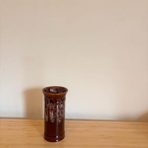 May include: A tall, cylindrical brown ceramic vase with a glossy finish. The vase features a dripped glaze design in shades of brown and cream. The vase is displayed on a wooden surface.