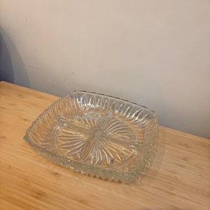 Op de afbeelding: Een heldere, vierkante glazen serveerschaal met drie compartimenten. De schaal heeft een gestructureerd, geribbeld ontwerp rond de randen en een radiaal patroon in het midden. Ideaal voor het serveren van snacks of hapjes.