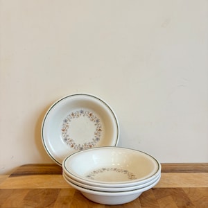 Può includere: Un set di quattro ciotole in ceramica bianca con un motivo floreale e un bordo verde. Le ciotole sono impilate su una superficie di legno. Il motivo floreale comprende fiori blu, arancioni e gialli.