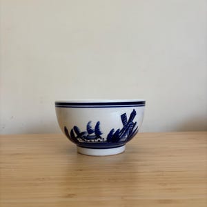 Könnte beinhalten: Eine weiße Keramikschale mit blauem Rand und blauem Landschaftsdesign mit Windmühlen und Bäumen. Die Schale hat eine abgerundete Form und steht auf einem kleinen Sockel. Das Design erinnert an Delfter Ware.