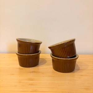 May include: Four brown ceramic ramekins with vertical ribbed detailing. The ramekins are stacked in pairs, with two sets of two. The interior of the ramekins is a lighter brown colour. They are arranged on a light brown wooden surface.