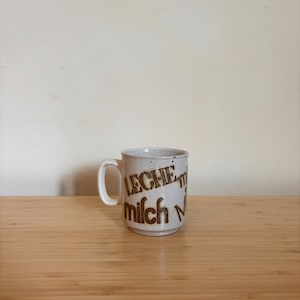 Könnte beinhalten: Eine weiße Keramiktasse mit brauner Beschriftung, die "LECHE-MILCH-LAIT" in einer Retro-Schriftart lautet.