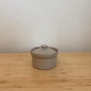 Könnte beinhalten: Eine kleine, gesprenkelte, beige Keramikdose mit Deckel. Die Dose steht auf einer Holzoberfläche.