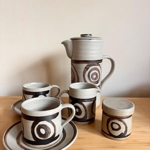 Könnte beinhalten: Ein Kaffee-Service aus Keramik in Creme- und Brauntönen. Das Set umfasst eine Kaffeekanne, einen Sahnekännchen, eine Zuckerdose und zwei Tassen mit Untertassen. Jedes Teil hat ein sich wiederholendes Kreisdesign.