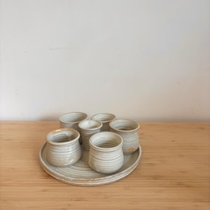 Könnte beinhalten: Ein Set aus sechs handgefertigten Keramikbechern und einem passenden Teller. Die Becher und der Teller sind hellbeige mit horizontalen strukturierten Linien. Die Becher sind unterschiedlich groß und stehen auf einem runden Teller.