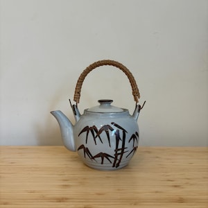 Peut inclure: Une théière en céramique grise avec un motif de bambou et une poignée tressée. La théière est posée sur une surface en bois.
