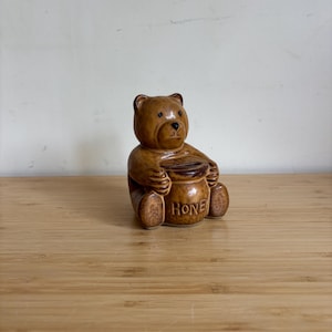 Può includere: Statuetta di orso in ceramica marrone che tiene un vasetto di miele. La parola "HONEY" è stampata sul vasetto. L'orso è seduto su una superficie di legno contro uno sfondo bianco.