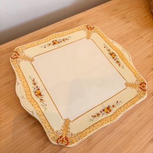 Könnte beinhalten: Ein quadratischer, cremefarbener Keramikservierteller mit floralem und geometrischem Design. Der Rand weist ein gelbes Punktmuster mit roten, orangefarbenen und gelben floralen Akzenten auf. Der Teller hat einen gewellten Rand und steht auf einer Holzoberfläche.
