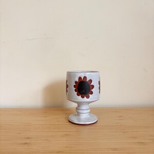 Könnte beinhalten: Ein Keramikbecher mit weiß gesprenkelter Oberfläche. Der Becher hat einen Sockel und ist mit sich wiederholenden braunen und schwarzen Sonnenblumenmotiven verziert. Der Rand und der Boden haben einen roten Akzent. Der Becher wird auf einer Holzoberfläche ausgestellt.