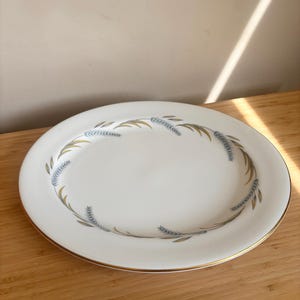 Könnte beinhalten: Ovale weiße Servierplatte mit goldenem Rand und einem Weizenähren-Design in Blau und Gold. Die Platte steht auf einer Holzoberfläche.