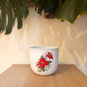Op de afbeelding: Een witte keramische plantenpot met een afgeronde vorm. De pot heeft een levendig bloemmotief van rode poinsettia's, groene bladeren en witte bloemen. De pot staat op een houten oppervlak.