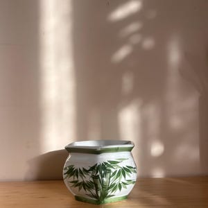 Könnte beinhalten: Weißer Keramiktopf mit grünem Bambus-Design. Der Topf hat einen quadratischen Boden und einen runden Körper.