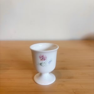 May include: White porcelain egg cup with a pedestal base. The cup features a delicate floral design in pink, green, and blue. The egg cup is designed with a flared rim and a rounded body.