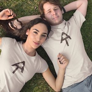 May include: Two people are lying on their backs on a grassy surface. They are wearing matching white t-shirts with a brown graphic of a stylized letter A with a heart inside. The woman is on the left and the man is on the right.