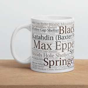 May include: White ceramic mug with a brown word cloud design featuring the names of shelters on the Appalachian Trail. The word cloud includes the names "Blood Mountain Shelter", "Carter Gap Shelter", "Whitley Gap Shelter", "Max Eppelsheimer Shelter", "Springer Shelter", "Low Gap Shelter", "Woods Hole Shelter", "Katahdin (Baxter Peak)", and "Black Mountain Shelter".