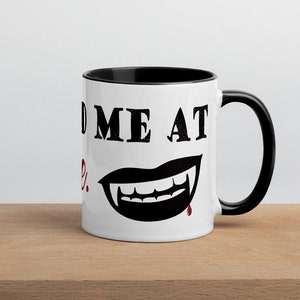 May include: White coffee mug with a black rim and handle. The mug has a black and red vampire mouth graphic and the text "Feed Me At Midnight".