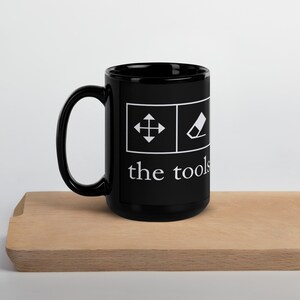 Op de afbeelding: Zwarte keramische mok met witte pictogrammen van een pijl en een gum, en de tekst "the tools" aan de voorkant.