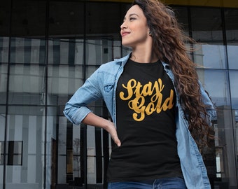May include: Black t-shirt with the words "Stay Gold" in gold script. The shirt is worn with a light blue denim jacket. The image is taken in front of a modern building with large glass windows.
