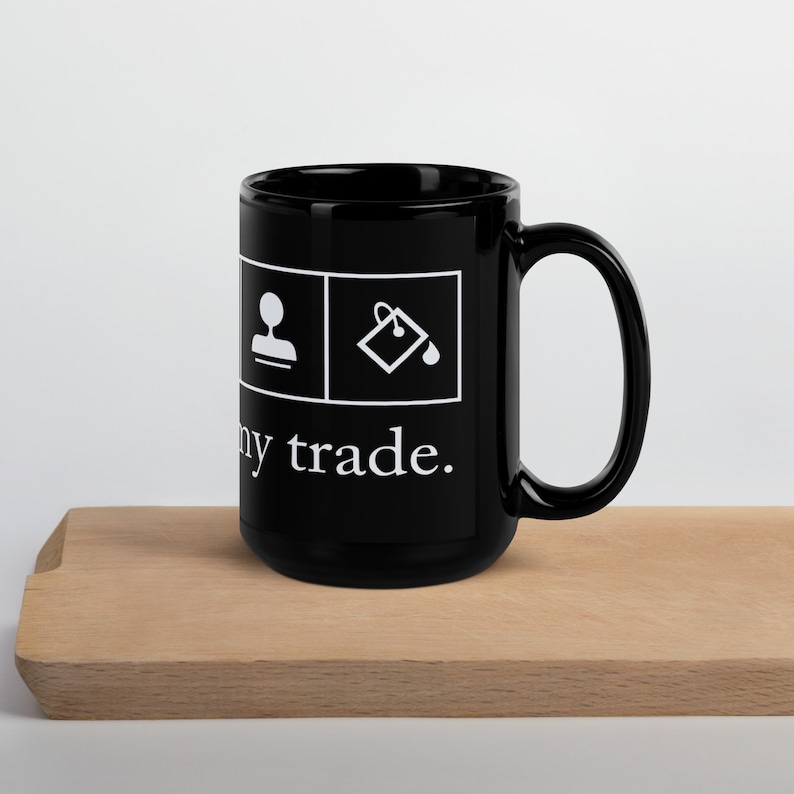 Op de afbeelding: Zwarte keramische mok met witte pictogrammen en de tekst "My trade." De pictogrammen tonen een persoon en een emmer verf.