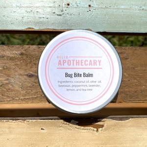 May include: A silver tin with a pink label that reads "HELLO APOTHECARY Bug Bite Balm" and lists the ingredients: coconut oil, olive oil, beeswax, peppermint, lavender, lemon, and tea tree.