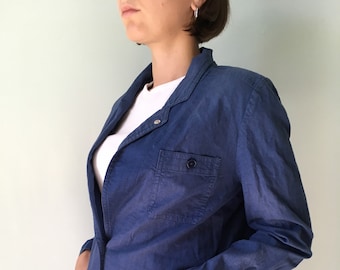 May include: A blue blazer with two front pockets and a button closure. The blazer has long sleeves and a collared neckline. The person is wearing a white t-shirt underneath. The blazer is a solid color and appears to be made of a soft fabric.