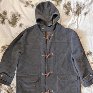 May include: A charcoal gray hooded duffel coat with toggle closures and two front pockets. The coat has a hood and long sleeves with buttoned cuffs. The toggles are brown leather and the buttons are a light brown color.