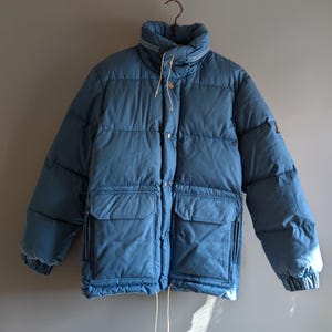 May include: A blue puffer jacket with a high collar and two front pockets. The jacket has a quilted design and is hanging from a metal hanger. The jacket has a drawstring at the bottom.