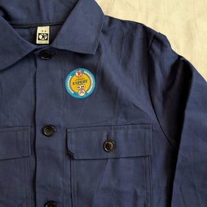 May include: A navy blue work shirt with a collar, two chest pockets, and black buttons. A circular patch on the chest reads "EXPERT" and "Gr. 44". The shirt is made of a durable fabric.