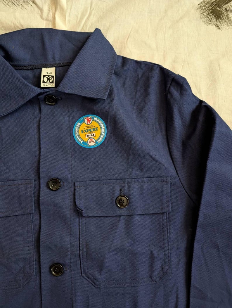May include: A navy blue work shirt with a collar, two chest pockets, and black buttons. A circular patch on the chest reads "EXPERT" and "Gr. 44". The shirt is made of a durable fabric.