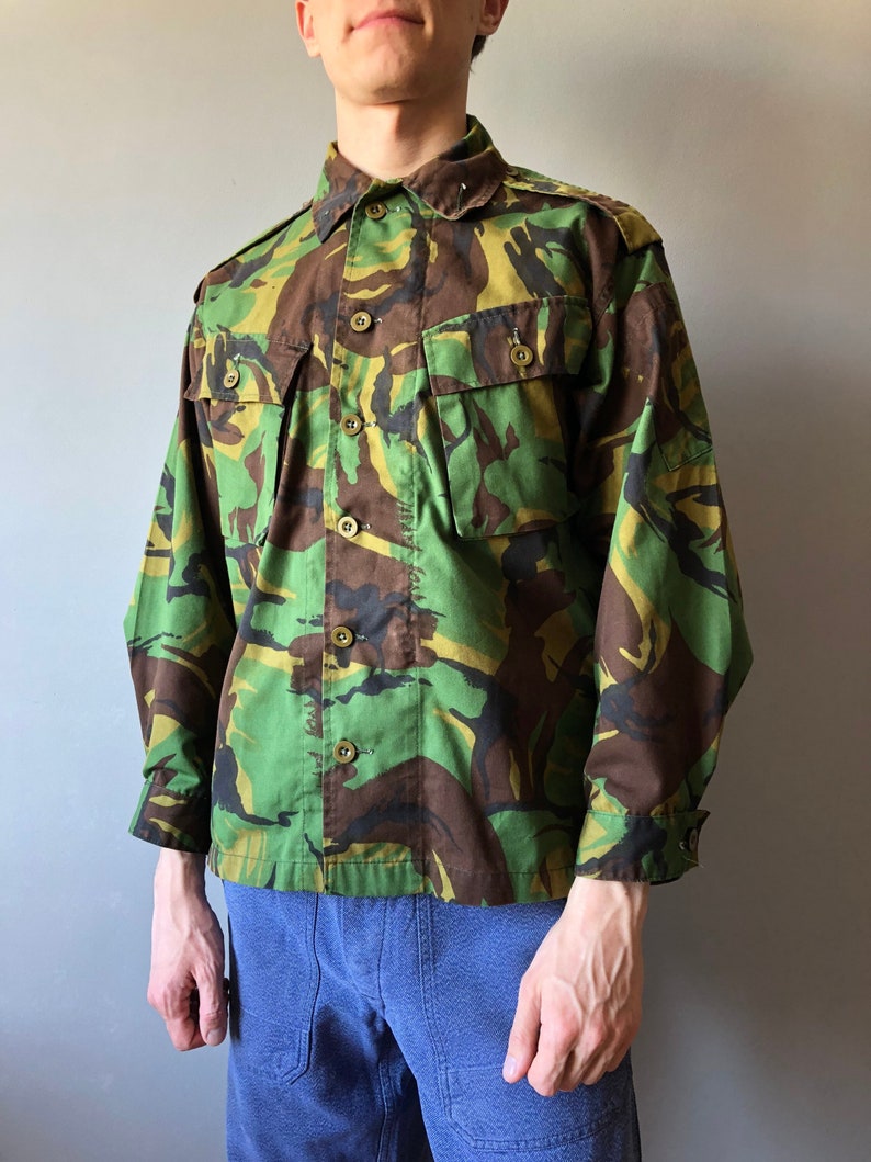 May include: A green, brown, and black camouflage button-up shirt with long sleeves and a button-down collar.