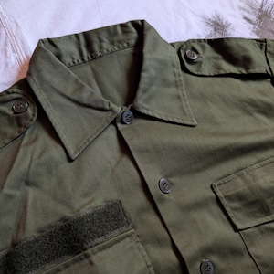 Può includere: Primo piano di una camicia verde oliva con bottoni. La camicia presenta un colletto, due tasche sul petto e una toppa in velcro sulla manica sinistra. Ha bottoni neri ed è realizzata in tessuto resistente.