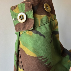 May include: Close-up of a camouflage fabric sleeve with two brown buttons. The fabric is a mix of green, brown, and yellow.