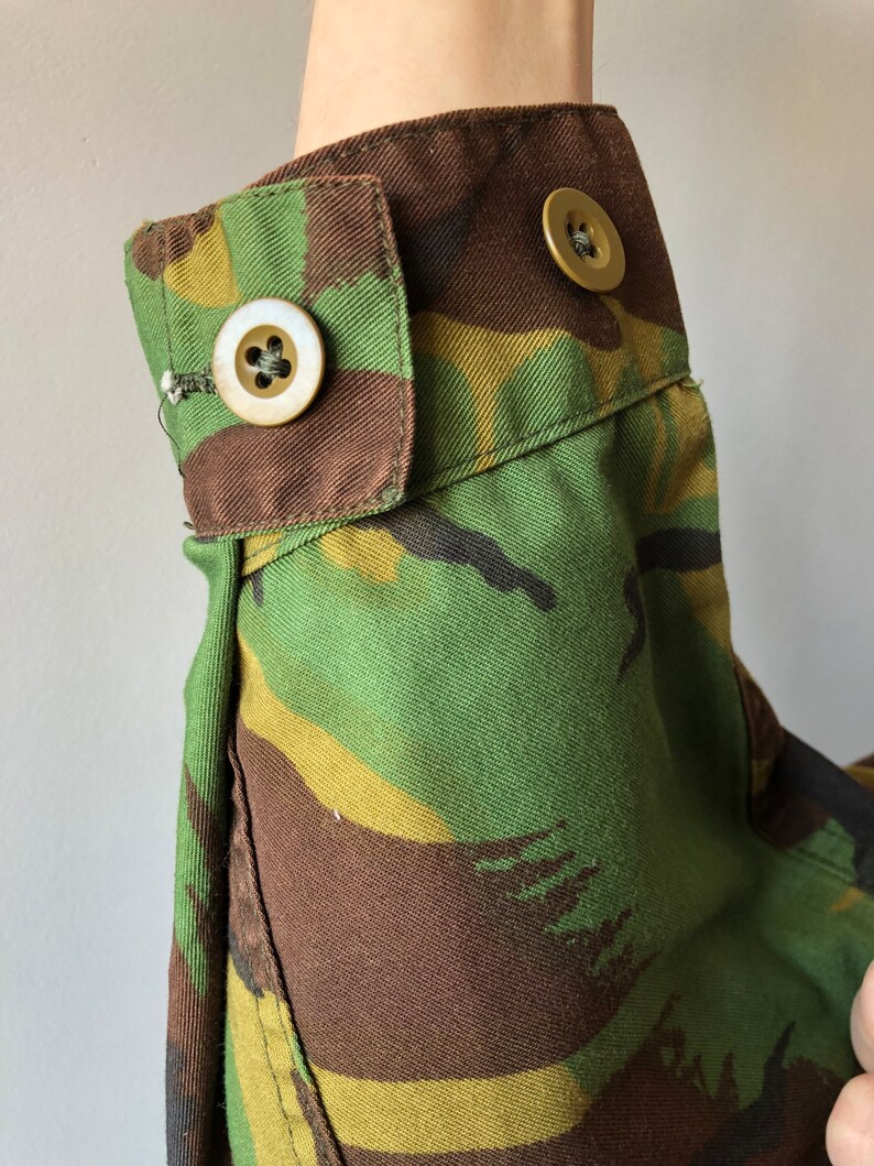 May include: Close-up of a camouflage fabric sleeve with two brown buttons. The fabric is a mix of green, brown, and yellow.