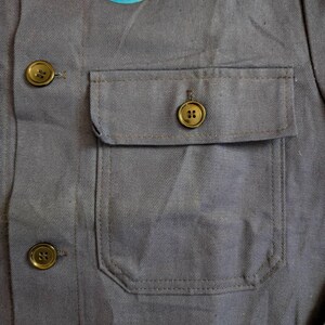 May include: Close-up of a gray work shirt with a chest pocket and three gold-colored buttons. A circular tag with the word "EXPERT" is attached to the shirt. The shirt is made of a durable fabric.