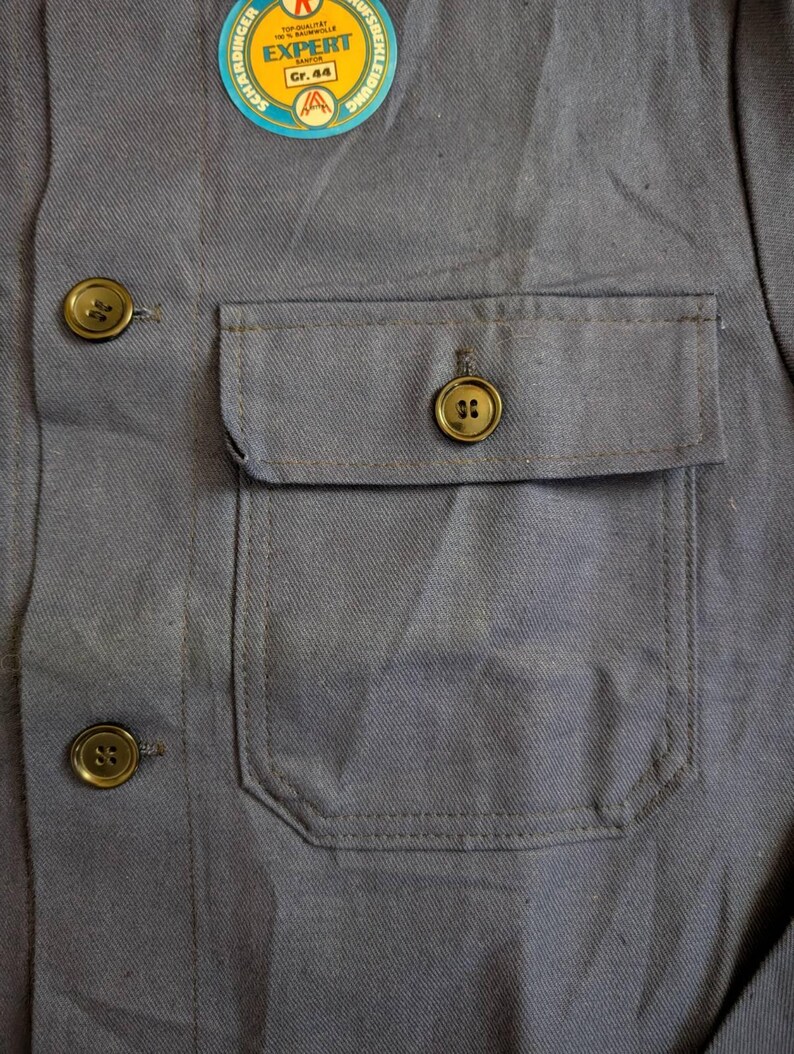 May include: Close-up of a gray work shirt with a chest pocket and three gold-colored buttons. A circular tag with the word "EXPERT" is attached to the shirt. The shirt is made of a durable fabric.