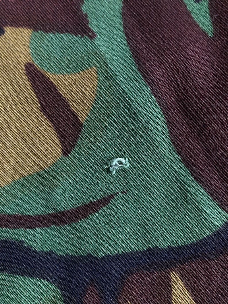 May include: Close-up of a green, brown, and black camouflage fabric with a small white thread loop.