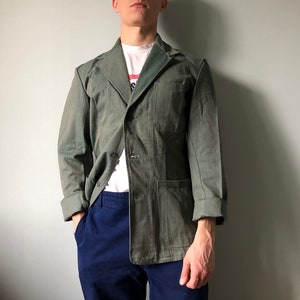 以下が含まれることがあります： ノッチカラー、ボタン留め、フロントポケットが特徴の、クラシックなデザインの緑色のジャケット。白いTシャツと青いパンツと組み合わせています。袖は折り返されており、裏地が見えています。