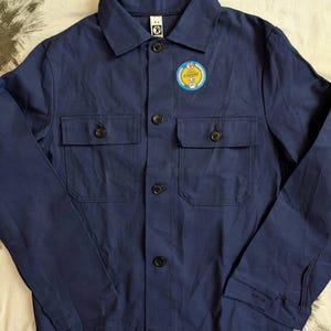 May include: A navy blue work jacket with two chest pockets and black buttons. A circular patch with the word "EXPERT" is on the left chest. The jacket has a classic collar and long sleeves.