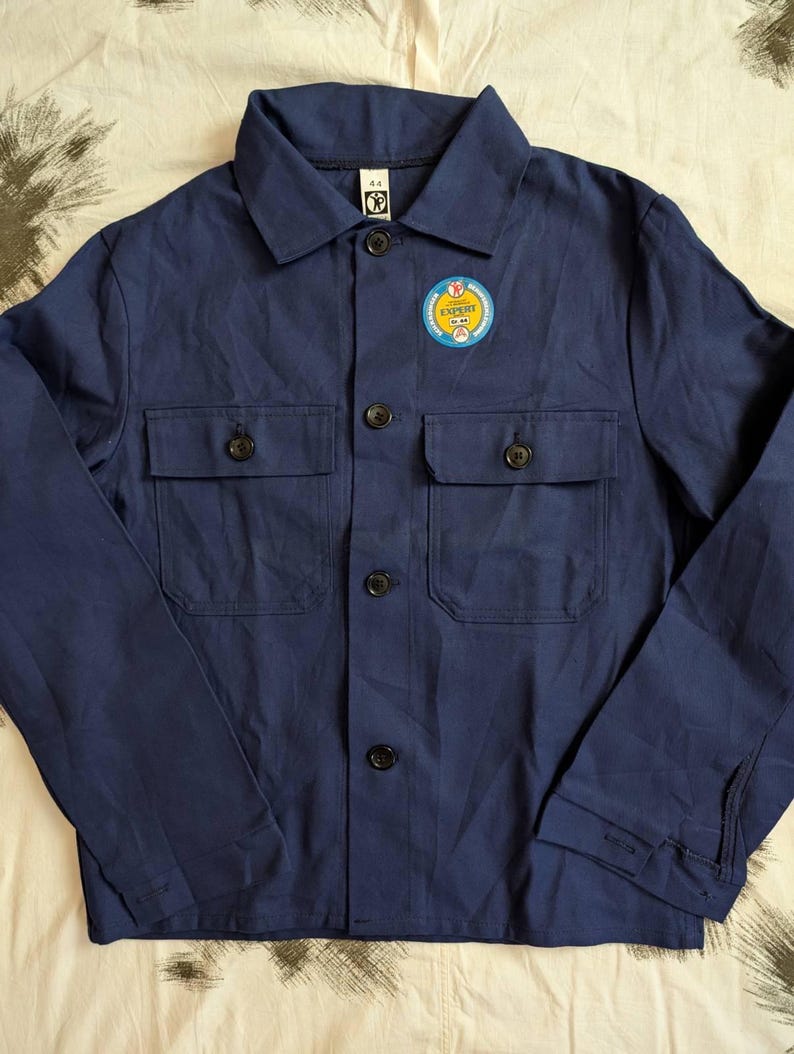 May include: A navy blue work jacket with two chest pockets and black buttons. A circular patch with the word "EXPERT" is on the left chest. The jacket has a classic collar and long sleeves.
