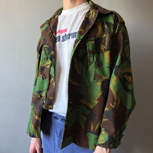 May include: A green, brown, and black camouflage jacket with a white t-shirt underneath. The t-shirt has the text "Original" and "en shrm" printed on it.