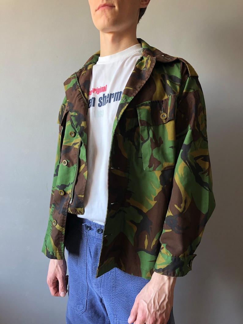 May include: A green, brown, and black camouflage jacket with a white t-shirt underneath. The t-shirt has the text "Original" and "en shrm" printed on it.
