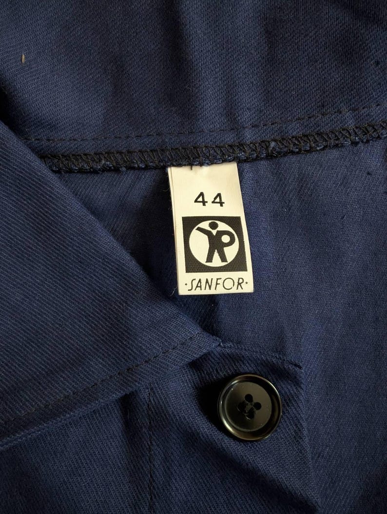 May include: Close-up of a navy blue work shirt with a black button and a label that reads "44" and "SANFOR". The label features a graphic of a person with outstretched arms.