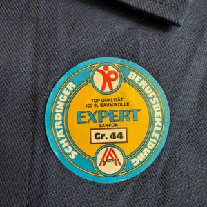 May include: A round, blue, yellow, and red sticker with the text "EXPERT SANFOR Gr. 44" and "100% BAUMWOLLE" on a blue fabric background. The sticker also features the text "SCHARDINGER" and "BERUFSBEKLEIDUNG".