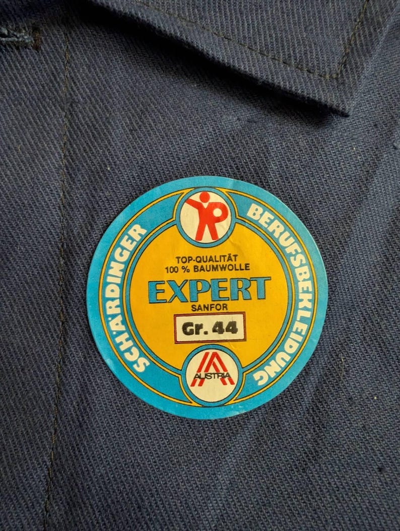 May include: A round, blue, yellow, and red sticker with the text "EXPERT SANFOR Gr. 44" and "100% BAUMWOLLE" on a blue fabric background. The sticker also features the text "SCHARDINGER" and "BERUFSBEKLEIDUNG".