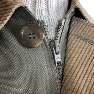 以下が含まれることがあります： コーデュロイの襟と大きな丸いボタンが付いた緑色のジャケットのクローズアップ。 ジャケットにはシルバーのジッパーが付いており、白いケーブルニットのセーターの上に重ね着されています。 ジッパープルには「OPTI」の文字が記されています。