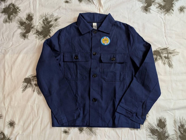 May include: A navy blue work jacket with a button-up front, two chest pockets, and a small patch on the upper left chest. The patch has a yellow and blue design with text. The jacket is made of a sturdy fabric.